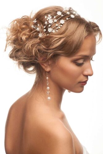 A woman with a curly updo hairstyle adorned with a delicate floral headpiece and pearl drop earrings, looking down gracefully.