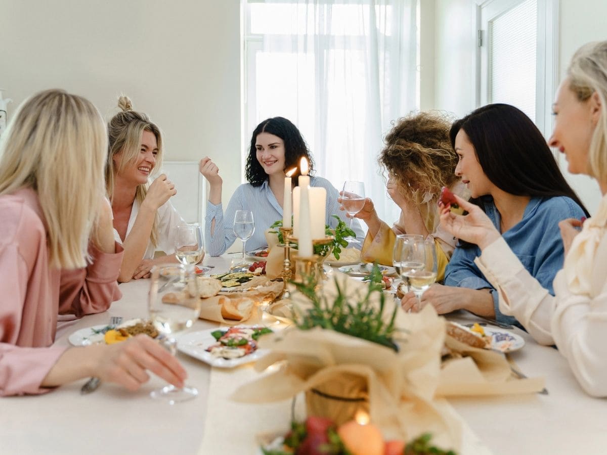 Young happy women friends drinking wine and laughing at the festive table served with fresh food and decorated with candles, avoiding bachelorette party drama.