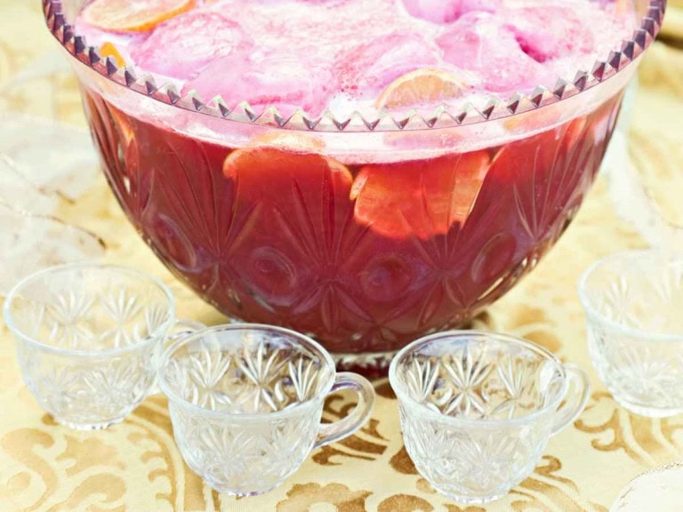 Bridal shower punch bowl filled with pink punch, citrus slices, and pink foam, surrounded by empty glass cups on a gold-patterned tablecloth.