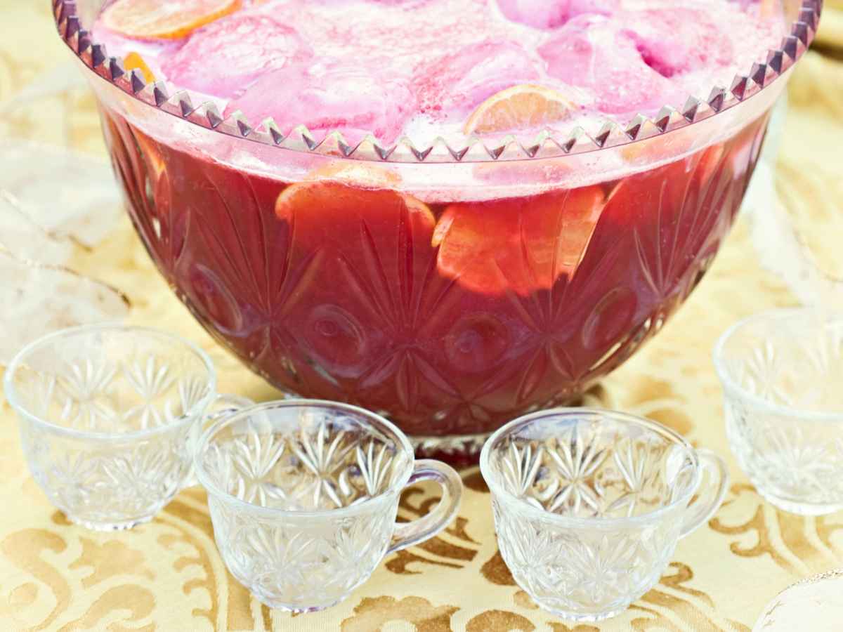 Bridal shower punch bowl filled with pink punch, citrus slices, and pink foam, surrounded by empty glass cups on a gold-patterned tablecloth.